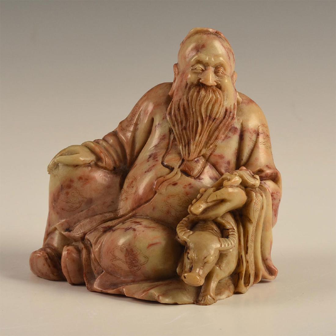 CHINESE CARVED SOAPSTONE FIGURAL SCHOLAR AND OX: Highly detailed and carved from a single block of soapstone, depicting an old recumbent Chinese scholar, possibly Lao Tzu, modeled with a small ox or water buffalo beneath the folds of his intricately
