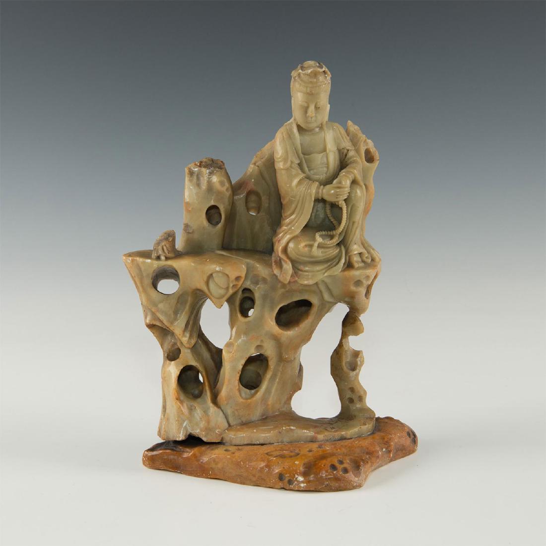 CHINESE CARVED SOAPSTONE GUANYIN ON ROCKY LEDGE: 20th century, detailed scene of figure resting on left knee on rocky edge. The figure is detailed with following robes and prayer beads. Guanyin is mythical and carries heavy symbolism in Chinese cult