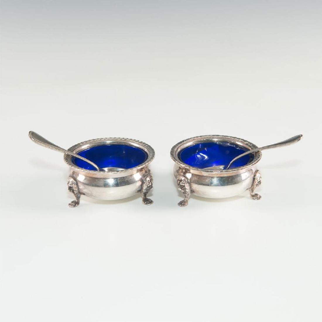 PAIR OF SILVER SALT CELLARS WITH SPOONS AND COBALT CUPS: Footed salts. Cobalt glass cups. Gadroon decoration to rims. A pair of three-footed silver salt cellars, with decorations of lions heads above the feet. Cobalt blue glass liners. Gadroon decoration to