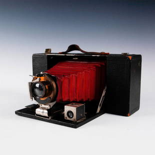 ANTIQUE KODAK TBI FPK AUTOMATIC FOLDING BELLOWS CAMERA: Black leatherette exterior. Maroon cordovan leather bellows. Bausch & Lomb lens incised with manufacturer stamp. Brown leather carry strap. Original merchants plate affixed ERNEST GOLDBACHER OPTICAL