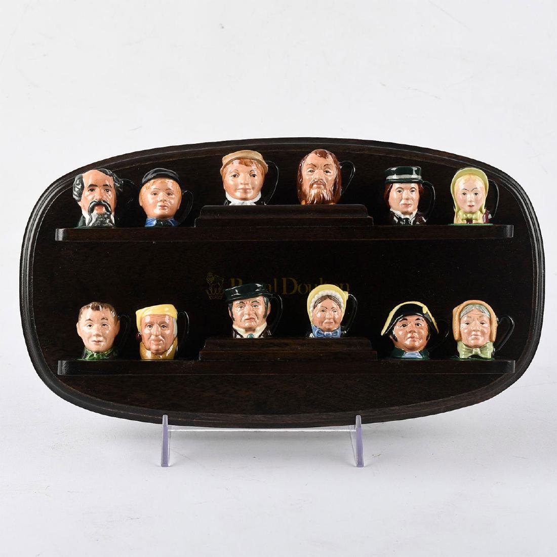 12 ROYAL DOULTON TINY CHARACTER JUGS AND DISPLAY STAND: Dickens commemorative set with Doulton stamped wooden stand. Characters include Bill Sykes D6684, Artful Dodger D6678, Charles Dickens D6676, Betsy Trotwood D6685, Little Nell D6681, Scrooge D6683, Ol