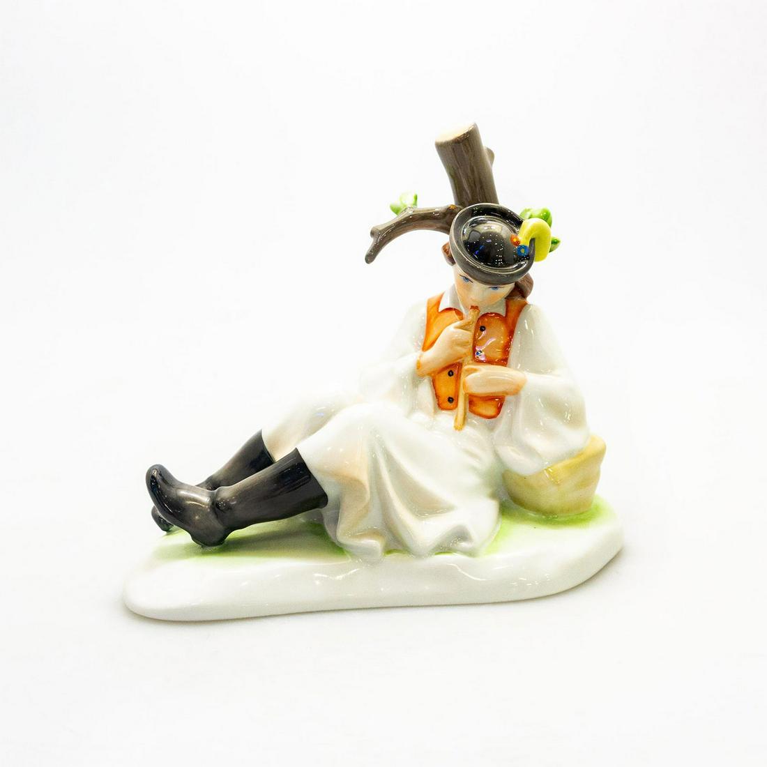 ZSOLNAY HUNAGRIAN PORCELAIN FIGURINE, MAN PLAYING FLUTE: Handpainted porcelain; man in hat and cloak seated beneath a tree stump playing a flute. Zsolnay backstamp. Dimensions: 7.5"H x 8.75"W Manufacturer: Zsolnay Country of Origin: Hungary