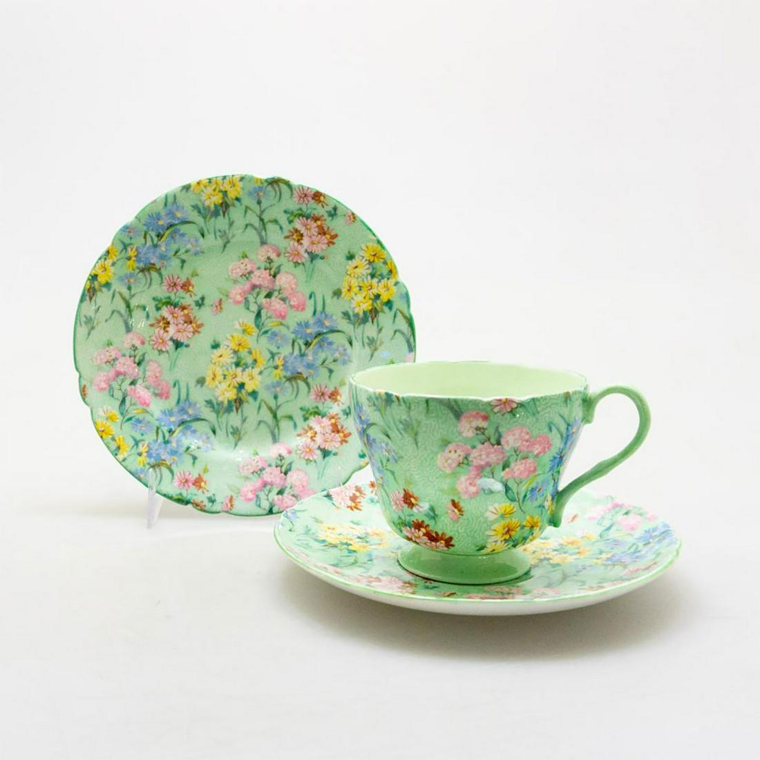 SHELLEY BONE CHINA ART DECO TEA TRIO, MELODY PATTERN: A glazed Art Deco trio with cup, saucer, and plate in seafoam green, profuse flora. Rich seafoam green background with pink, blue, yellow, and red daisy floral decoration. SHELLEY FINE BONE CHINA ENGL
