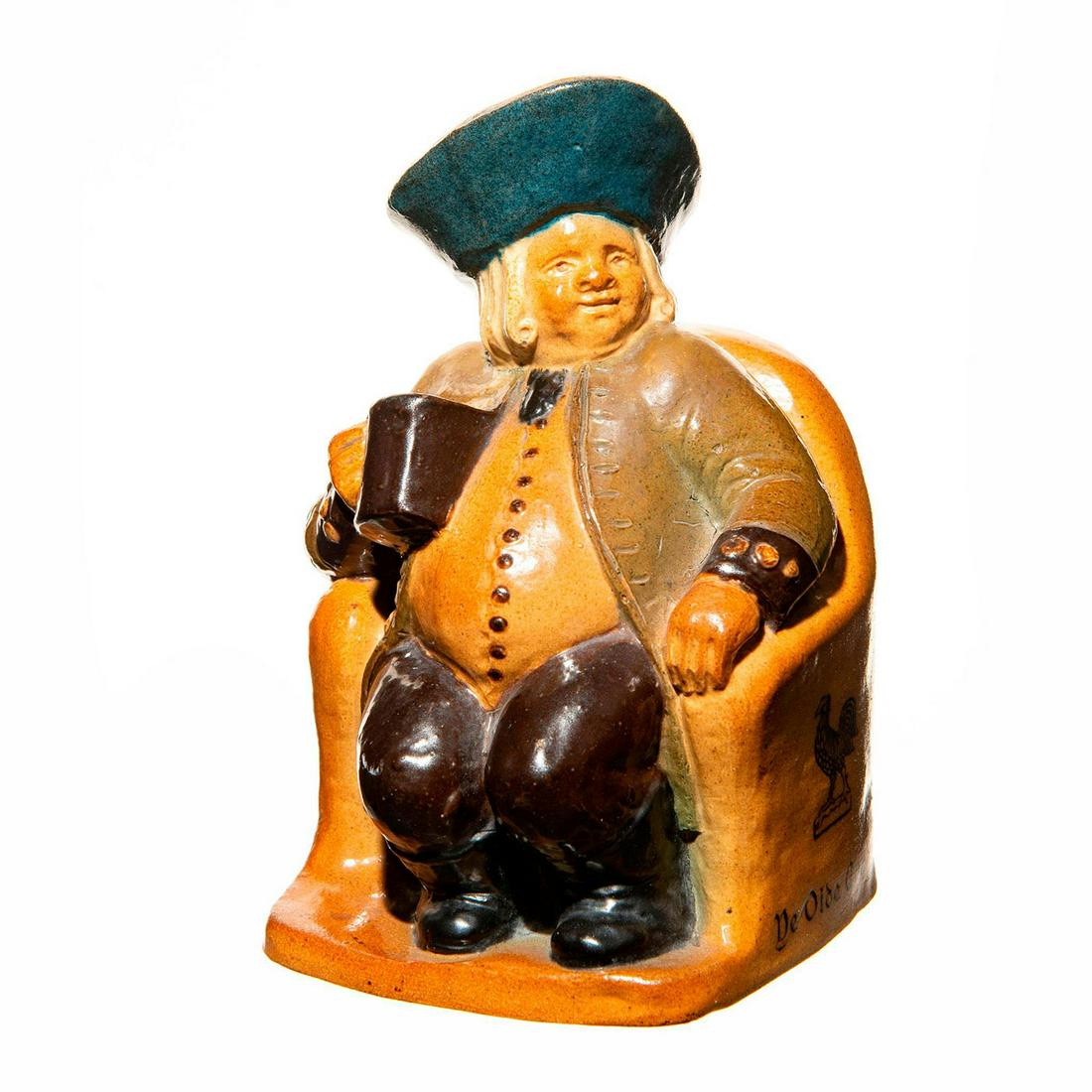 Doulton Lambeth Seated Toby Man: Toby man sitting down on large light brown chair. He is dressed in an olive jacket and a light orange vest with black buttons. He is also wearing a navy hat and holding a black mug. The chair reads; Y