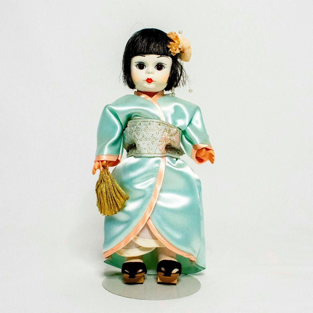 Madame Alexander Doll, Japan: Doll with golden brown eyes, black hair, and Geisha makeup. Wearing a light blue kimono. She wears white socks, okobo sandals, and carries a gold fan. Eyes close when tilted. Metal doll stand included