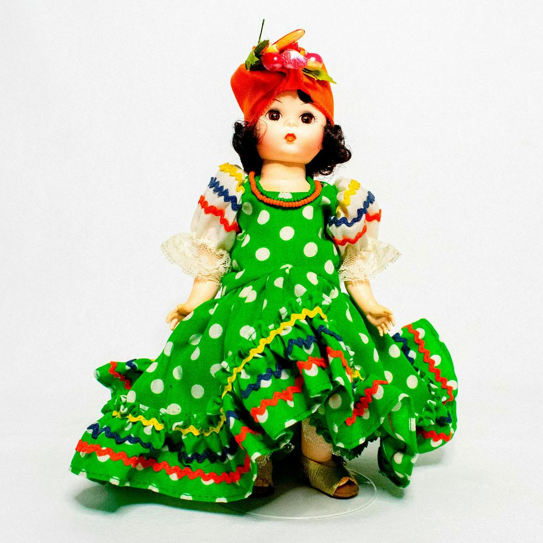 Madame Alexander Doll, Brazil: Doll with golden brown eyes and dark hair. She wears a green polka dot dress with zig zag ruffled trim. Her accessories include gold sandals, green and orange beaded necklaces, and an orange headdress