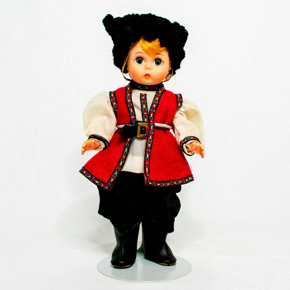 Madame Alexander Doll, Cossack: Doll with blue-grey eyes and blond hair. Wears black pants, white shirt, and red vest with embroidered trim. Black boots, a belt, and a furry black hat completes the look. Metal doll stand included. #