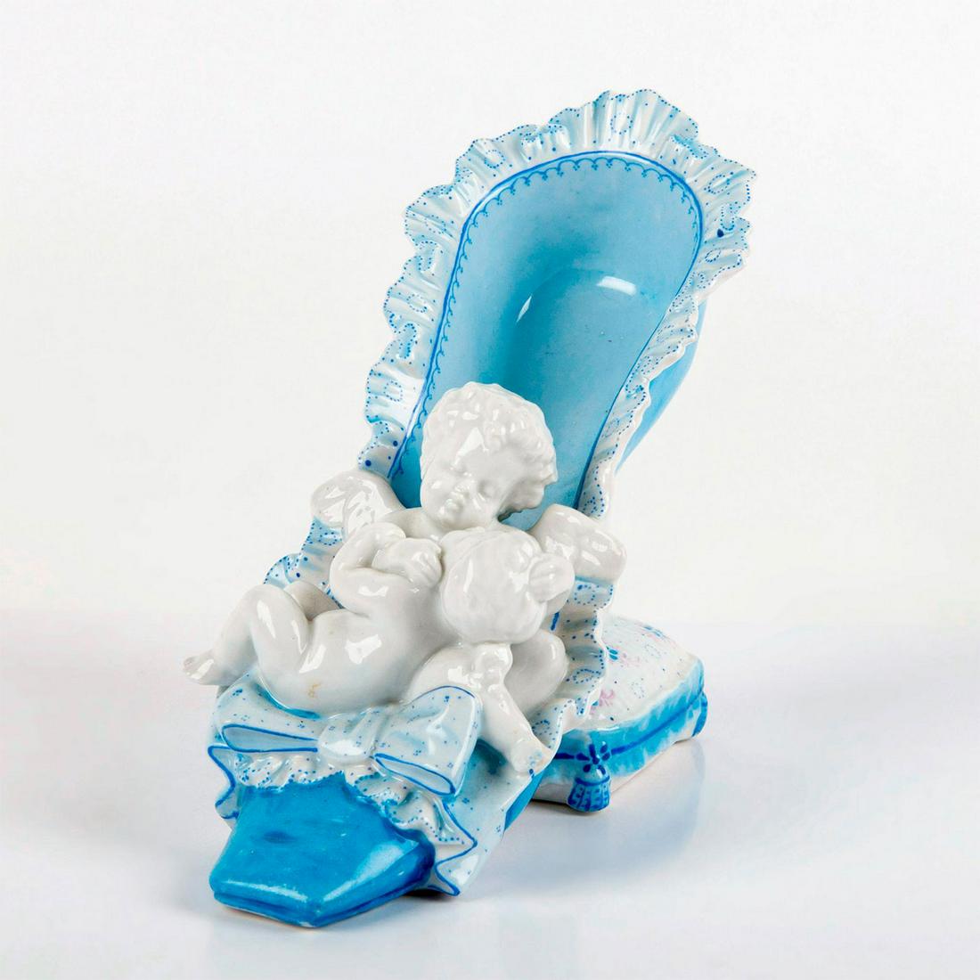 Von Schierholz Porcelain Figurine, Angel In Shoe: Blue and white ruffled slipper on a pillow stool. In the front a little angle holding a baby. Glazed finish. Depose Impressed on the bottom. Blue crossed lines mark. Minimal wear and tear due to age.