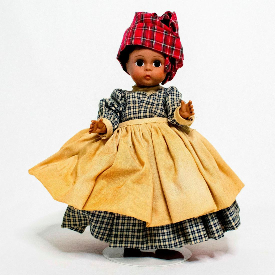 Madame Alexander Doll, Mammy: Doll with dark eyes and short dark hair. She wears a blue and beige plaid dress, with beige apron. She has a red skirt beneath her dress to add volume, white pantaloons, and black socks. With a red pl