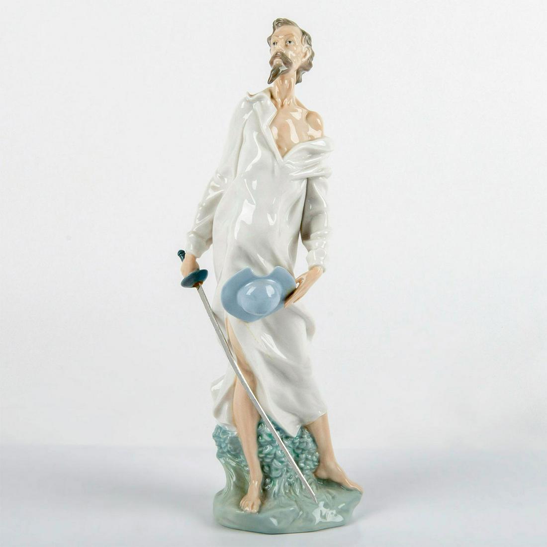Nao By Lladro Porcelain Figurine, Don Quixote: Glazed; Standing tall in white nightdress. Sword in one hand, blue hat in the other hand. Nao backstamp. Issued: 20th c. Dimensions: 12"H Manufacturer: Lladro Country of Origin: Spain
