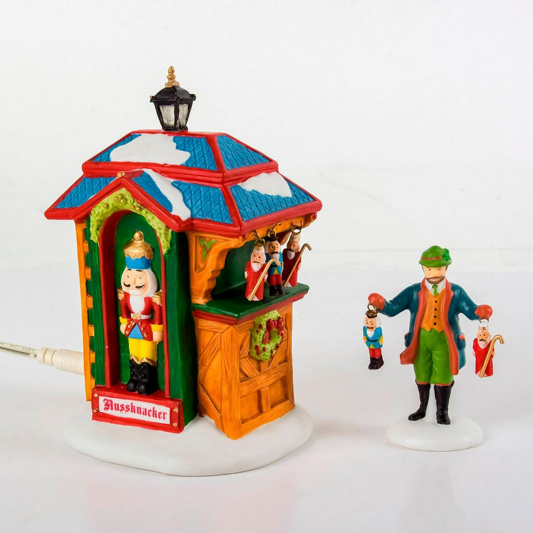 Set Of 2 Department 56 Figures, Nutcracker Booth: Handpainted porcelain; lighted nutcracker booth and salesman peddling his wares. Alpine Village Series. With original box. Department 56 backstamps. Comes with battery pack; batteries not included. No