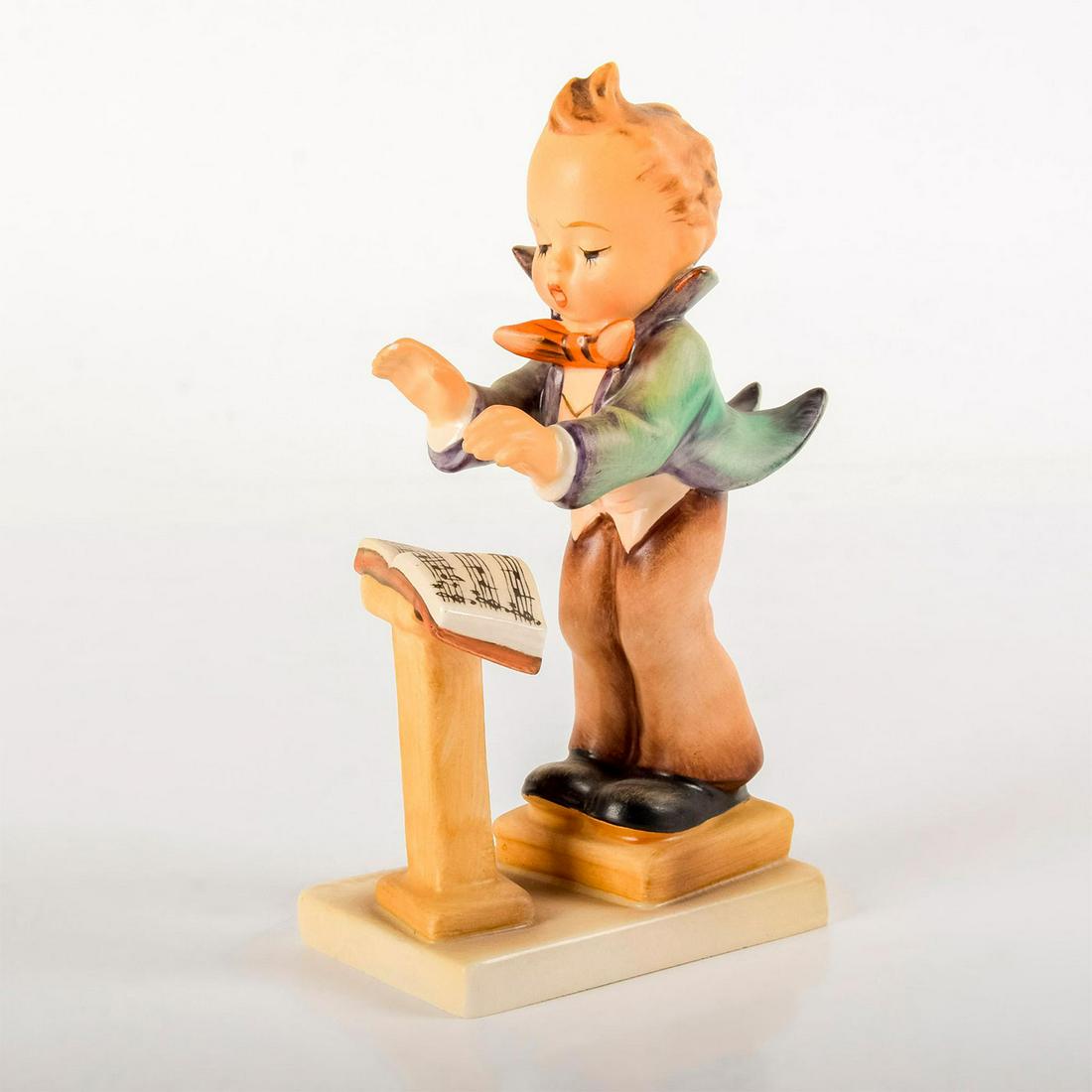 Band Leader 129 - Goebel Hummel Figurine: Matte porcelain, hand painted. Band conductor in green coat reading a sheet of music. 'M. I. Hummel' inscribed on base. Goebel backstamp. Age related crazing. Minor chip on right hand. Weight: