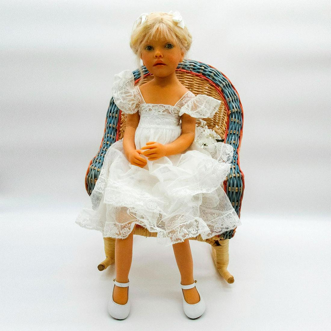 Christening Doll, Camille: Limited edition doll in satin white lace dress and faux leather shoes. Cotton body sitting in a wicker rocking chair. Camille written on the body. Signed. Age related wear and yellow eyes. Re-glued le