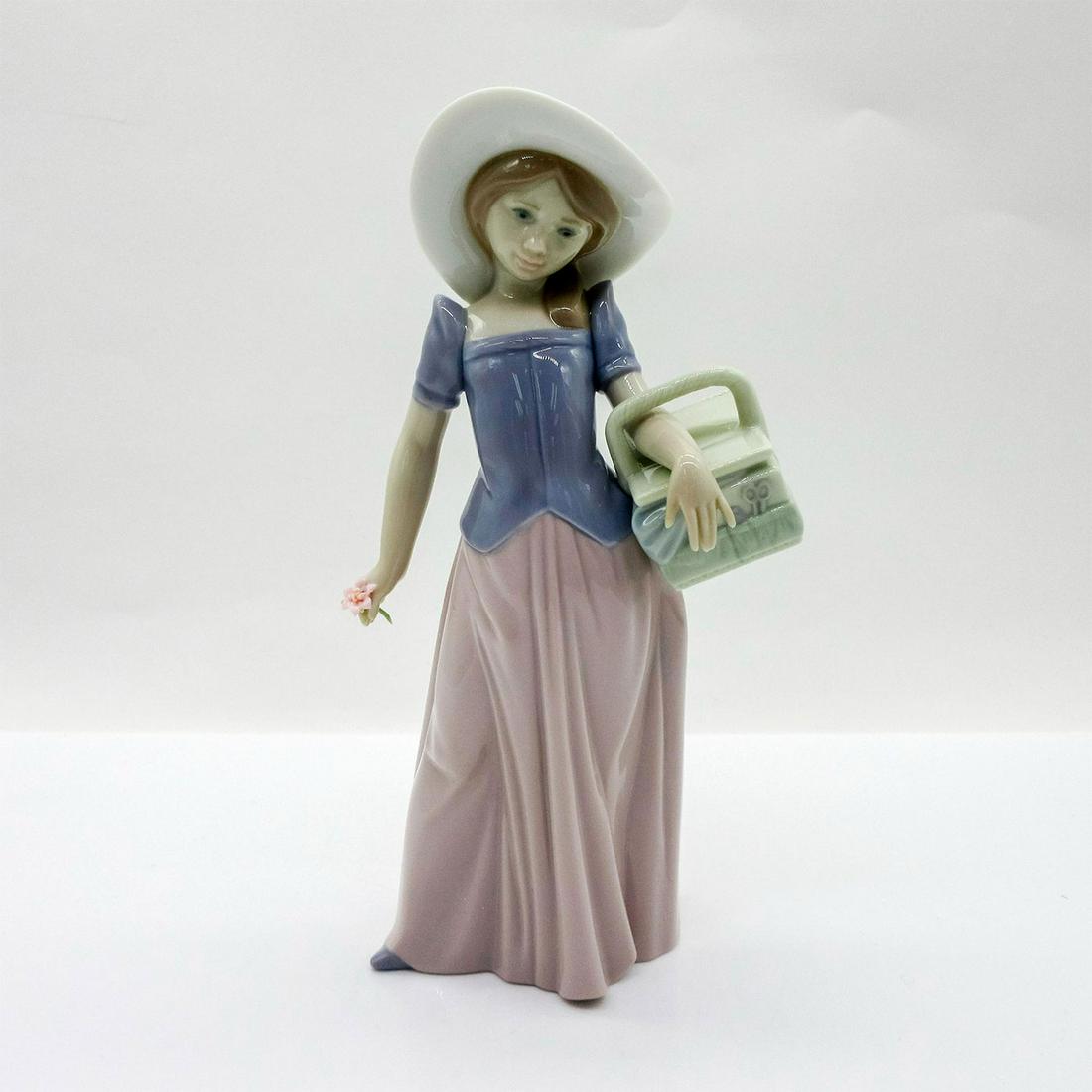 Tailor Made 1006489 - Lladro Porcelain Figurine: Glazed figure of a woman in a pink skirt, purple blouse and sun hat holding a basket and pink flower. Lladro backstamp. In original box, 5"H x 11"W x 5.5"L. Artist: Marco Antonio Nogueron Issued: 1997