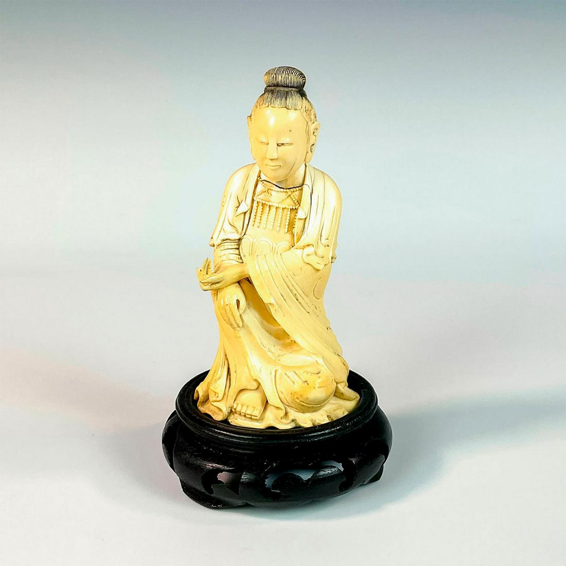 Chinese Carved Bone Seated Guanyin Figurine with Base: A figure of the Bodhisattva in flowing robes, holding her palm facing up. This item has a base included. Figurine: 2.5in L x 2.25in H x 5in W. Base: 3.5in W x 1.25in H. Country of Origin: China