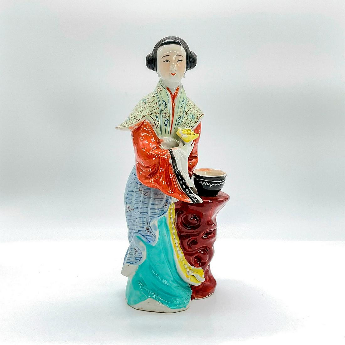 Chinese Figurine, Lady Holding Flower: Vintage glazed ceramic figure of a standing lady in traditional red, pale green and blue robe with Guang sleeves, turquoise skirt, holding a yellow flower. She leans on a slim round table with a bowl