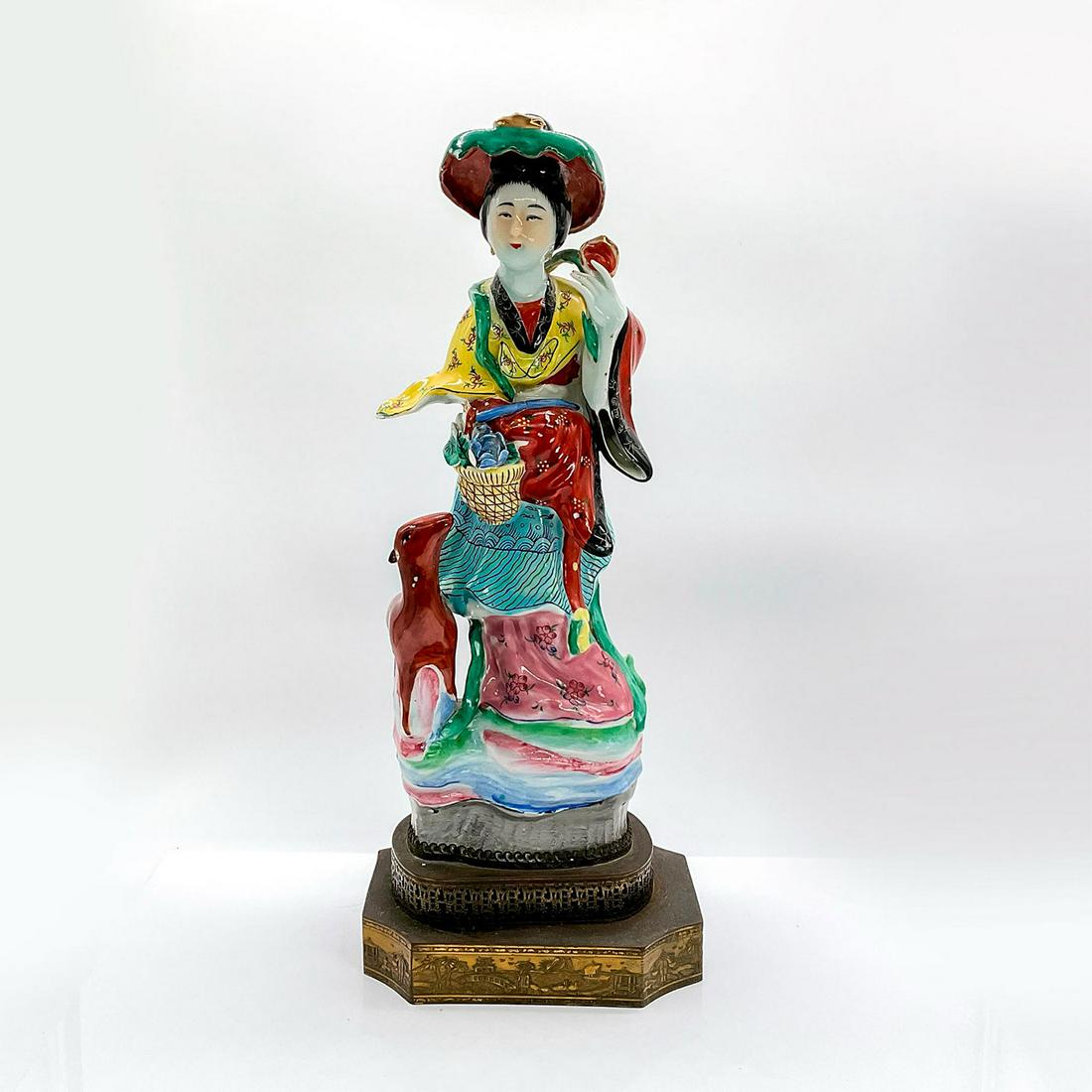 Vintage Chinese Figurine, He Xiangu With Lamb: Ceramic glazed figure of one of the Eight Immortals in a colorful robe and green hat holding a basket of blue flowers with a brown small deer next to her. This figure is attached to a brass base stand