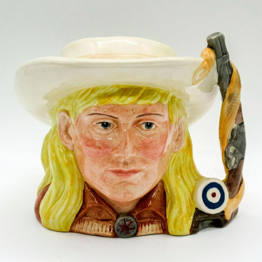 Annie Oakley D6732 - Odd Size - Royal Doulton Character Jug: Yellow hair; cream hat; brown tunic. Phoebe Anne Moses (1860 - 1926) learned to shoot at the age of eight and helped support her family by killing game for a hotel in Cincinnati, Ohio. At 15, she defe