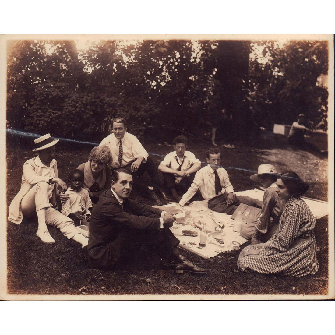 Original Monochrome Photograph, Picnic Meet: Featuring group of people collected together outside for picnic lunch setting on the grass. Presumably a film still or scene from movie released sometime between late 19th to early 20th century. Issue