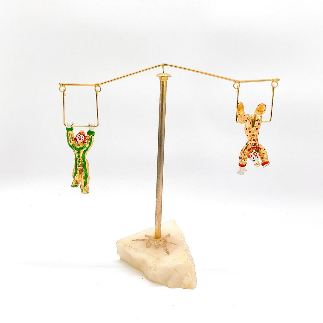 Swinging Trapeze Clown Sculpture: Two clown performers balance on trapeze frames. Metal frame and figures painted gold, green and red. Stone base. In the style of artist Ron Lee. Issued: 20th c. Dimensions: 10"L x 5.5"W x 10"H