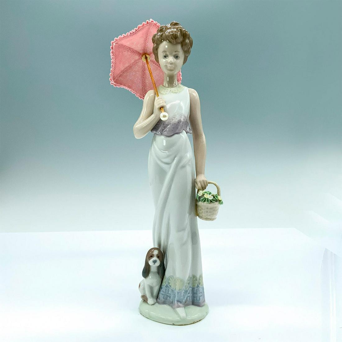 Garden Classic 01007617 - Lladro Porcelain Figurine: Depicts a woman in a white sun dress carrying a basket of flowers alongside her dog while holding a parasol. Lladro backstamp. Artist: Juan Huerta Issued: 1991 Dimensions: 2.25"L x 2.25"W x 9.5"H Manu