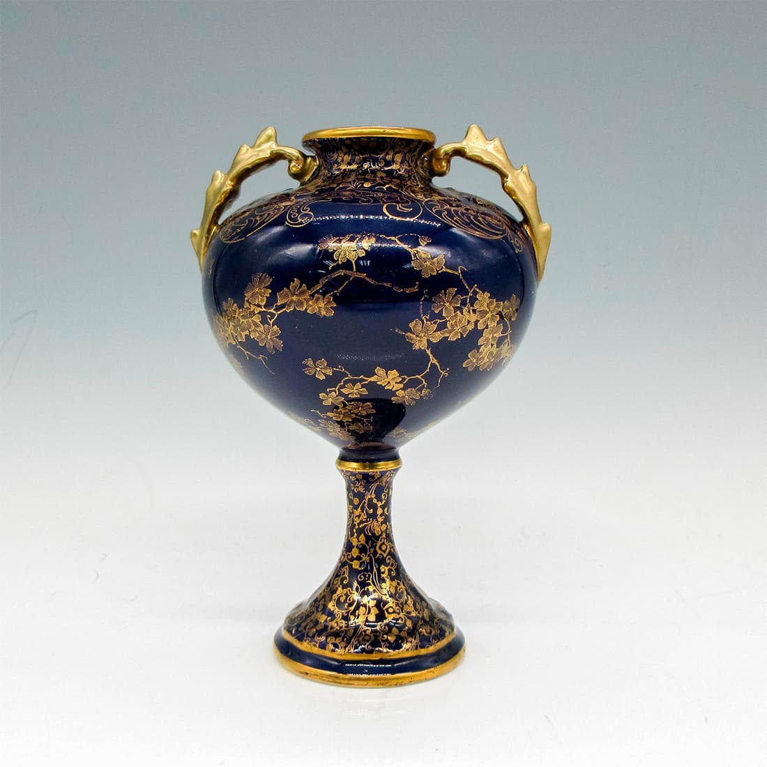 Royal Doulton Porcelain Cobalt Blue Pedestal Vase: Tapered vase with raised gilded design of leaves and vines. Gilded banding at neck and pedestal. Gold toned twin handles. Royal Doulton backstamp. Artist: Robert Allen Issued: c.1900 Dimensions: 4"L x