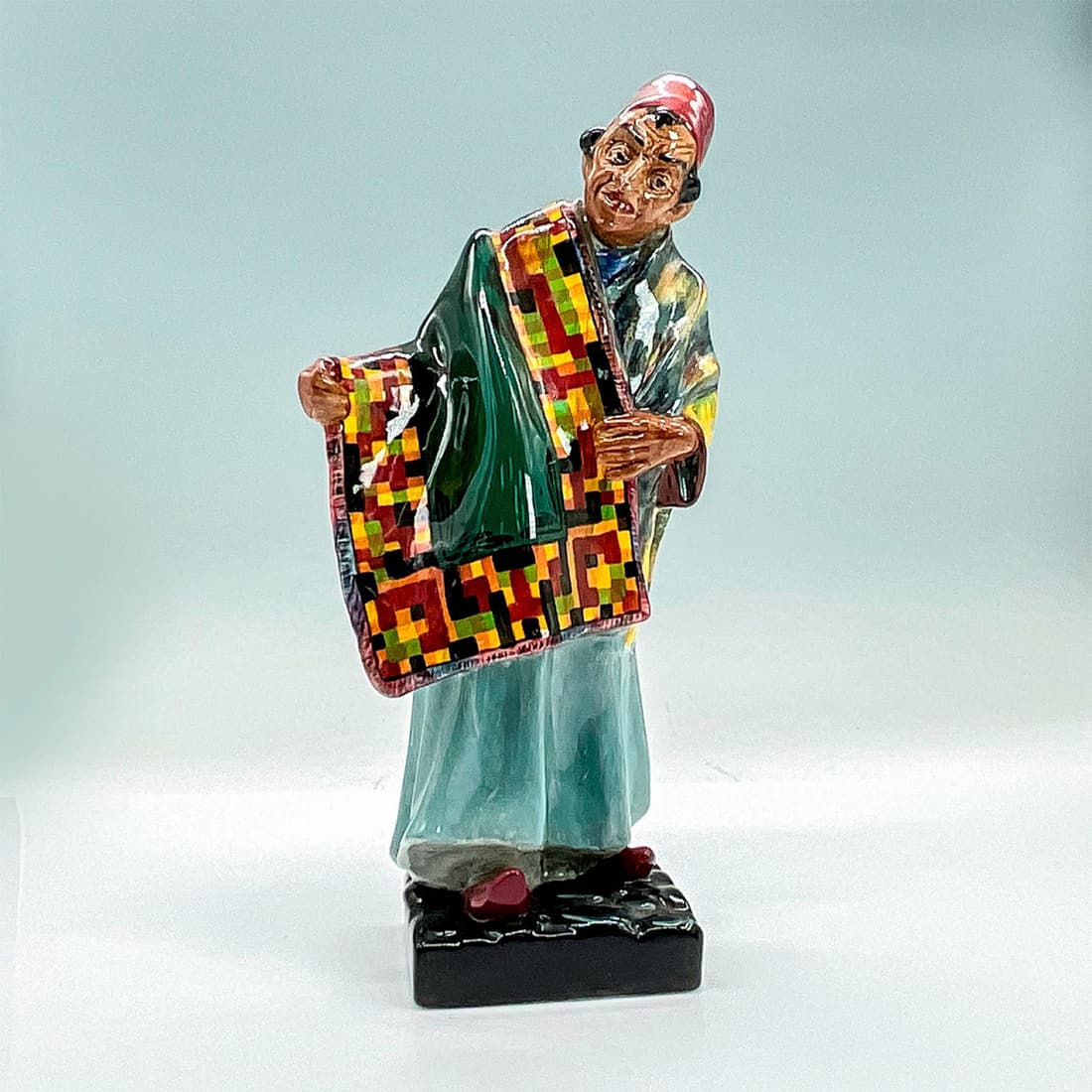 Carpet Seller HN1464 - Royal Doulton Figurine: He stands displaying a multi-colored rug, ready to make a deal. Royal Doulton backstamp. Part of the Middle Eastern Heritage Series. Artist: Leslie Harradine Issued: c. 1969 Dimensions: 4.25"L x 3.25"