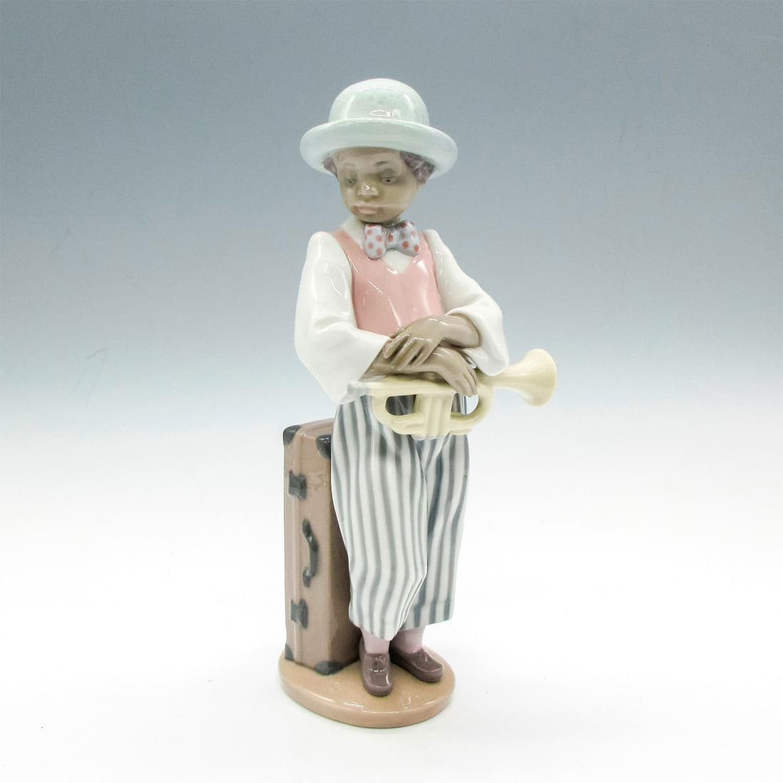 Lladro Porcelain Figurine, Jazz Horn 1005832: Glazed figural of boy with horn. Dressed in stripe pants with polka dot bow tie. Lladro backstamp. This item has its original box: 5.75"L x 11"W x 5"H. Artist: Rafael Lozano Issued: 1991-2017 Dimensio