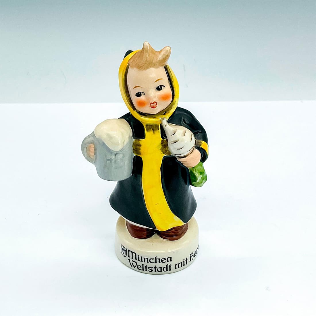 Goebel Hummel Figurine, Munchen Weltstadt Mit Herz: A boy in a black and yellow robe is carrying a mug of beer in one hand and turnip in the other hand. Goebel backstamp and LMun 44 imprint. Issued: 1964 - 1972 Dimensions: 2"L x 2.25"W x 5.25"H Manufac