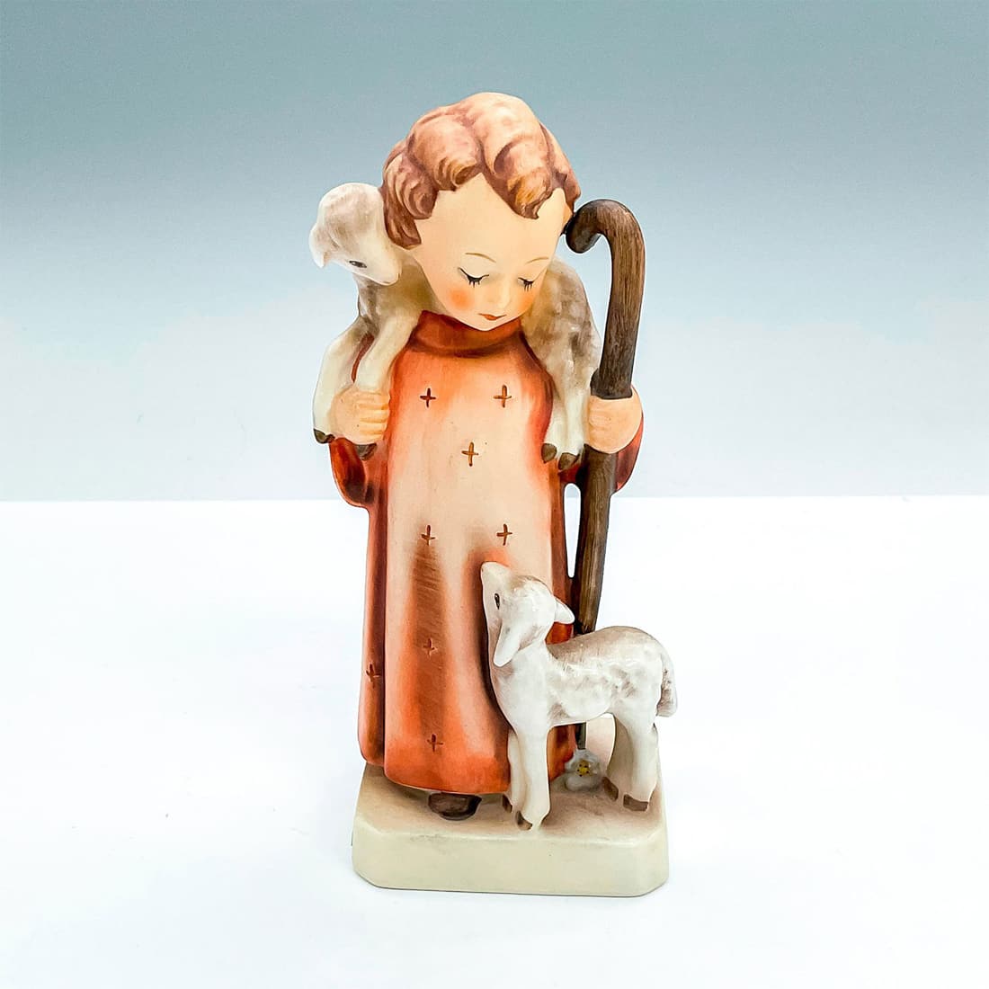 Goebel Hummel Figurine, Good Shepherd: Matte porcelain, hand painted. Young girl in red robe standing with two lambs. 'M. I. Hummel' inscribed on base. Goebel backstamp. Issued: 1979 - 1990 Dimensions: 2.75"L x 2.25"W x 6.5"H Manufacturer: