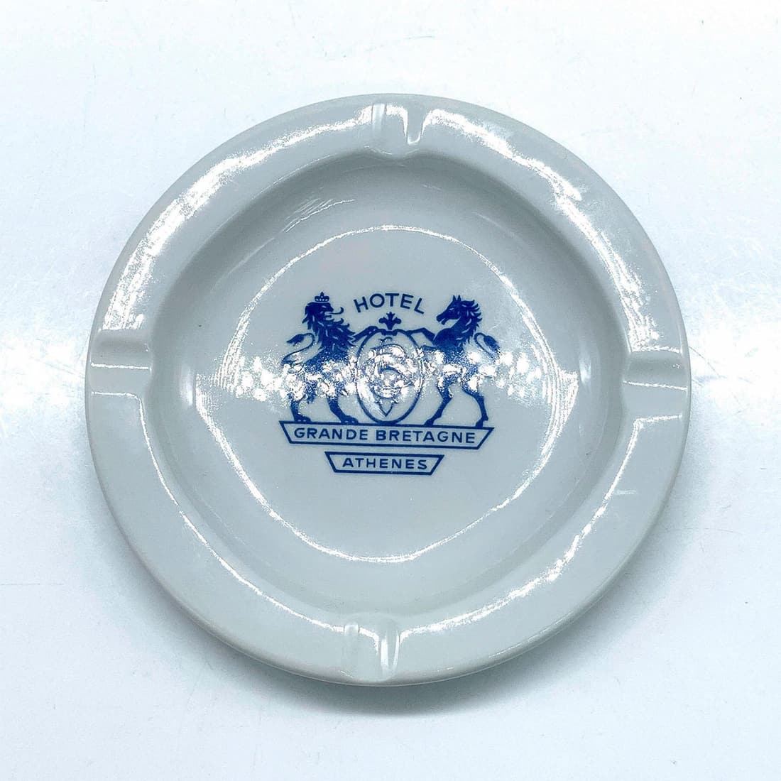 Hotel Grande Bretagne Souvenir Ashtray: Collectible porcelain ashtray from the Bretagne Grande Hotel in Athens. Blue emblem on front with 4 cigarette spots. Ionia Greece backstamp. Dimensions: 5.5" dia. Country of Origin: Greece