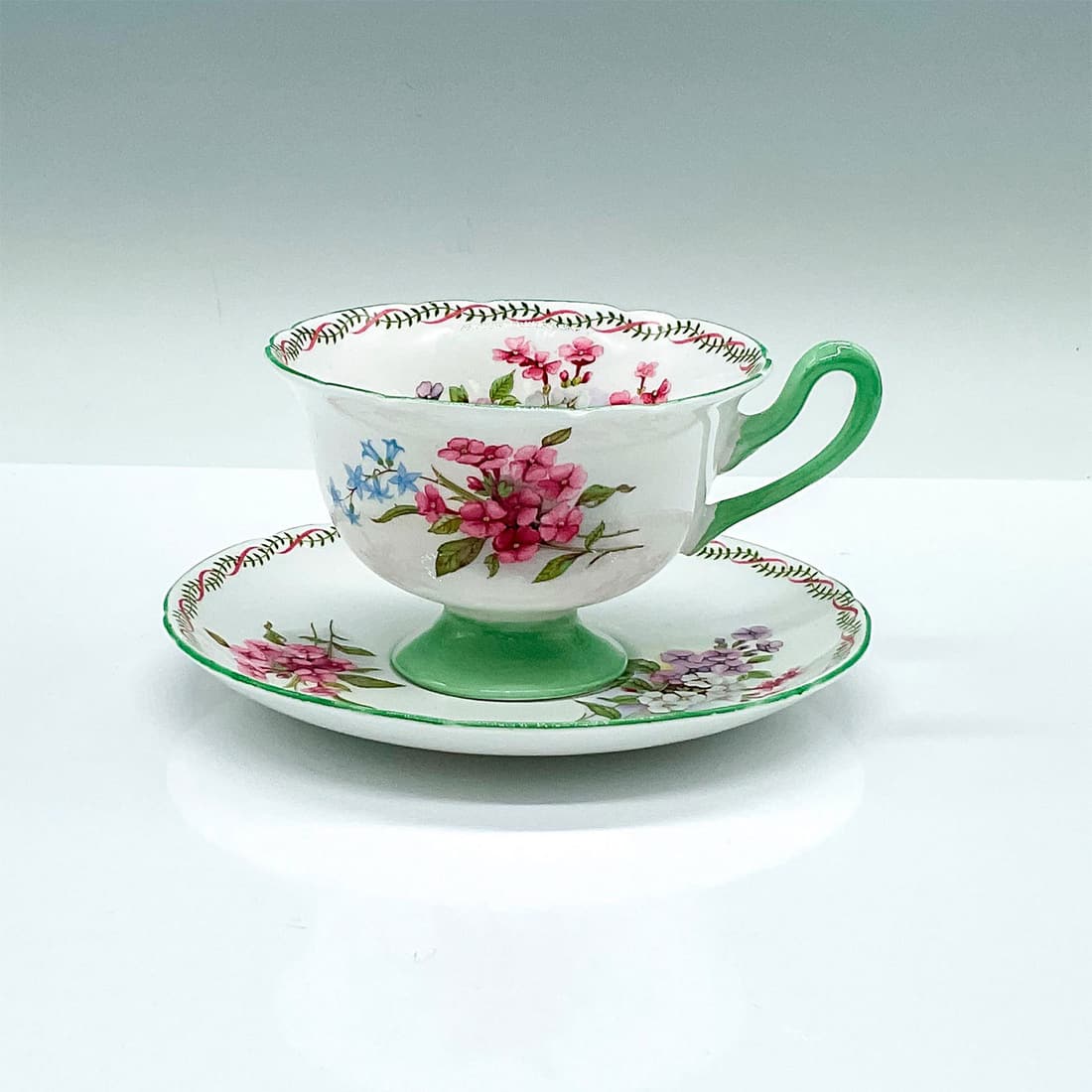 Vintage Shelley Fine Bone China Cup and Saucer: White ground tea cup and saucer set with green trim and ivy pink ribbon on the rim. Decorated with wildflowers. Shelley backstamps and marked 13512. Cup: 4.75"L x 4"W x 2.5"H. Saucer: 0.75"H x 5.5"H.