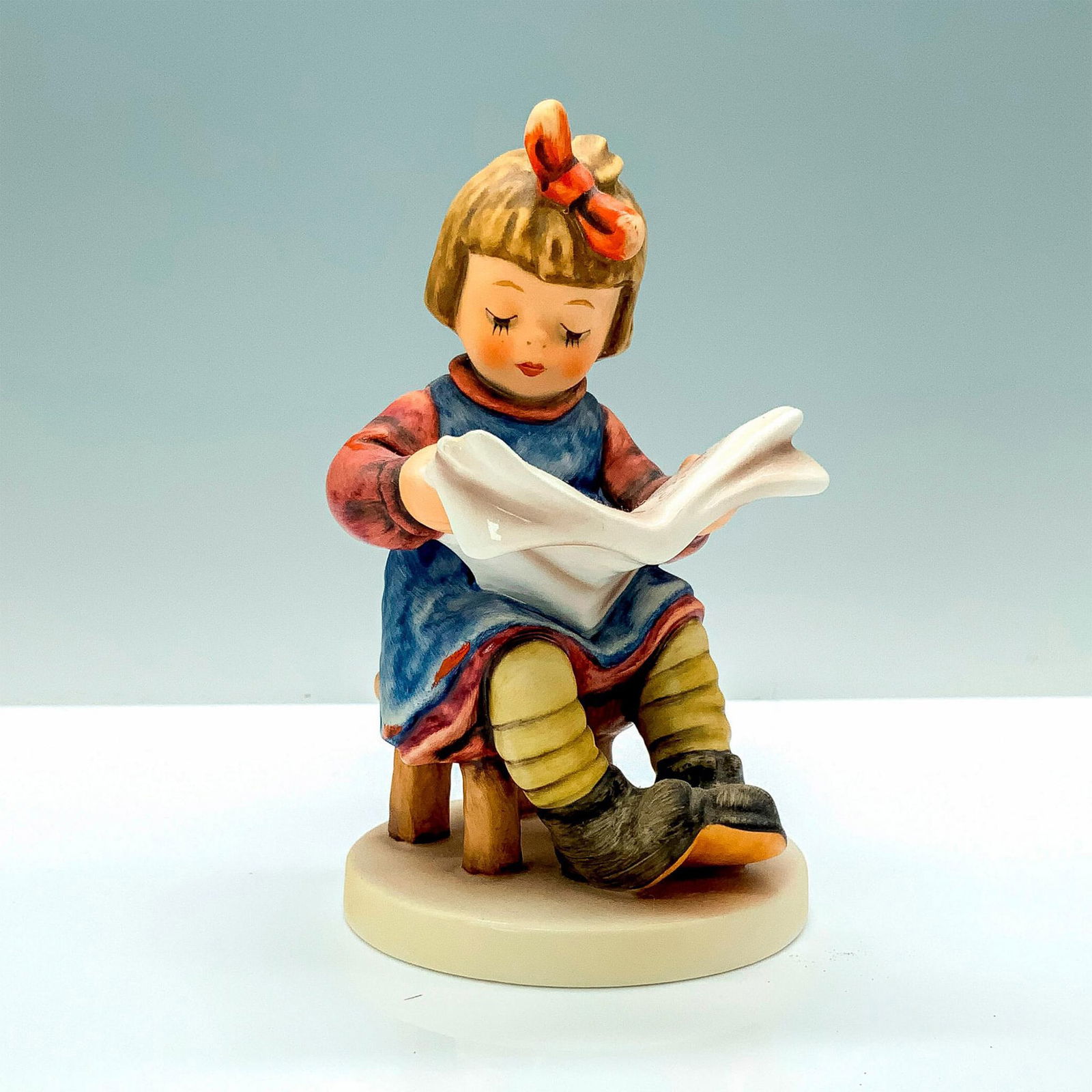 Goebel Hummel Figurine, What's News?: A figure of a young girl in a red dress and blue apron sitting on a wooden stool reading the newspaper. Goebel Hummel backstamp. Issued: 1989 Dimensions: 3"L x 3"W x 5"H Manufacturer: Goebel Hummel Co