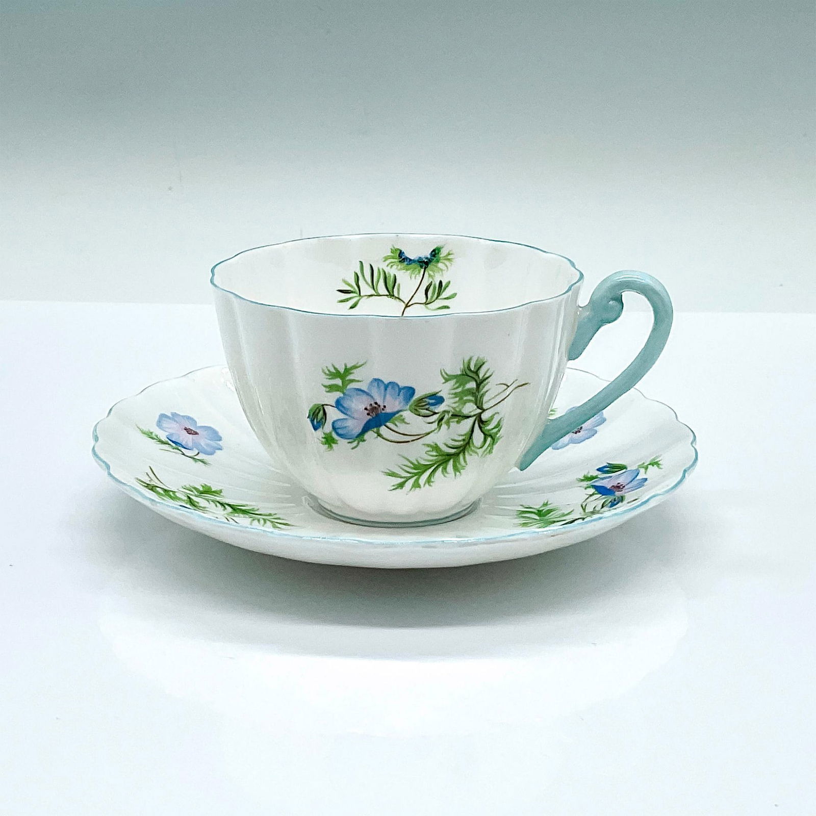 Shelley Bone China Tea Cup and Saucer, Blue Poppy: Tea cup and saucer set, depicting hand painted blue flowers. Light blue trim. Shelley backstamps. Cup: 4.25"L x 3.5"W x 2.25"H. Saucer: 5.75"W x 1"H. Pattern number: 14168. Issued: 20th century Manufa