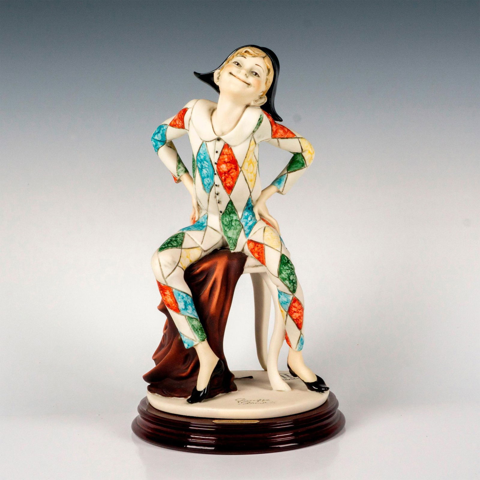 Giuseppe Armani Sculpture, Arlecchino Harlequin: A hand painted figure depicting a young boy sitting on a bench in a harlequin costume. Sculpture is attached on a round wooden base. Giuseppe Armani signature and backstamp. Issued: 1991 Dimensions: 6