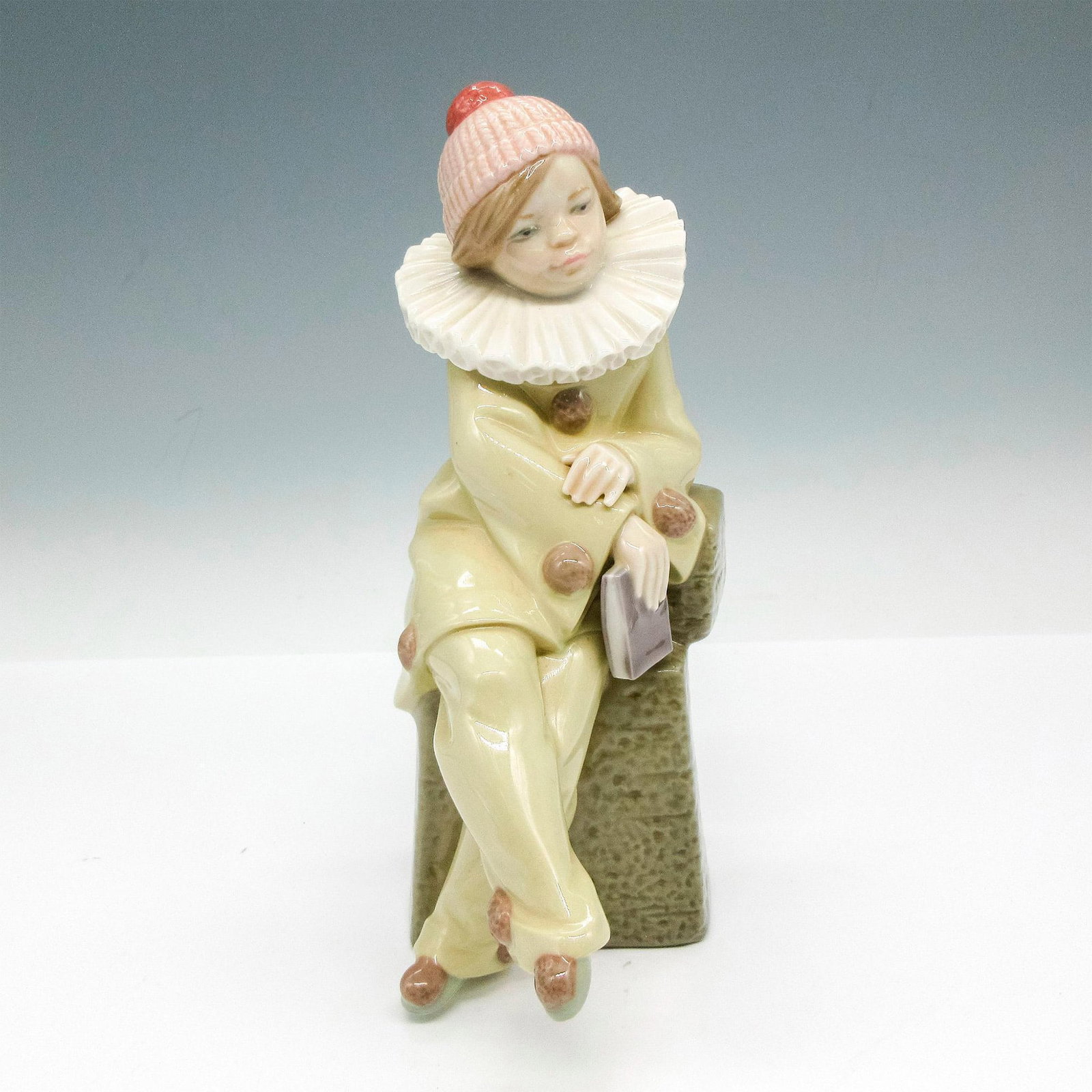 Lladro Porcelain Figurine, Little Jester 1005203: Glazed figural seated on base, dressed in decorated outfit with ruffled collar. Lladro backstamp. Artist: Juan Huerta Issued: 1984 - 1993 Dimensions: 3"L x 5"W x 8.5"H Manufacturer: Lladro Country of