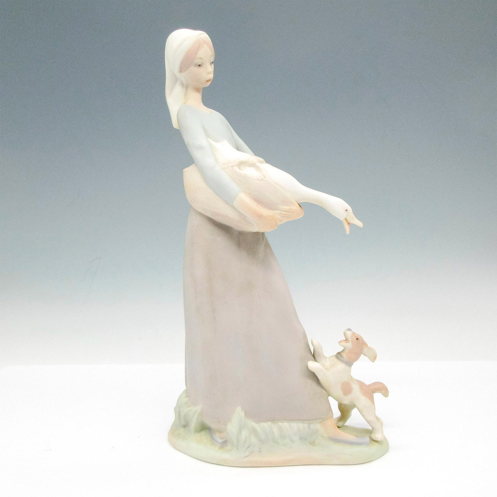 Nao by Lladro Porcelain Figurine, Girl with Goose and Dog: Matte finish on standing figural colored in soft cream, tan and blue. NAO backstamp. Issued: 20th century Dimensions: 5.25"L x 3"W x 10"H Manufacturer: Nao by Lladro Country of Origin: Spain