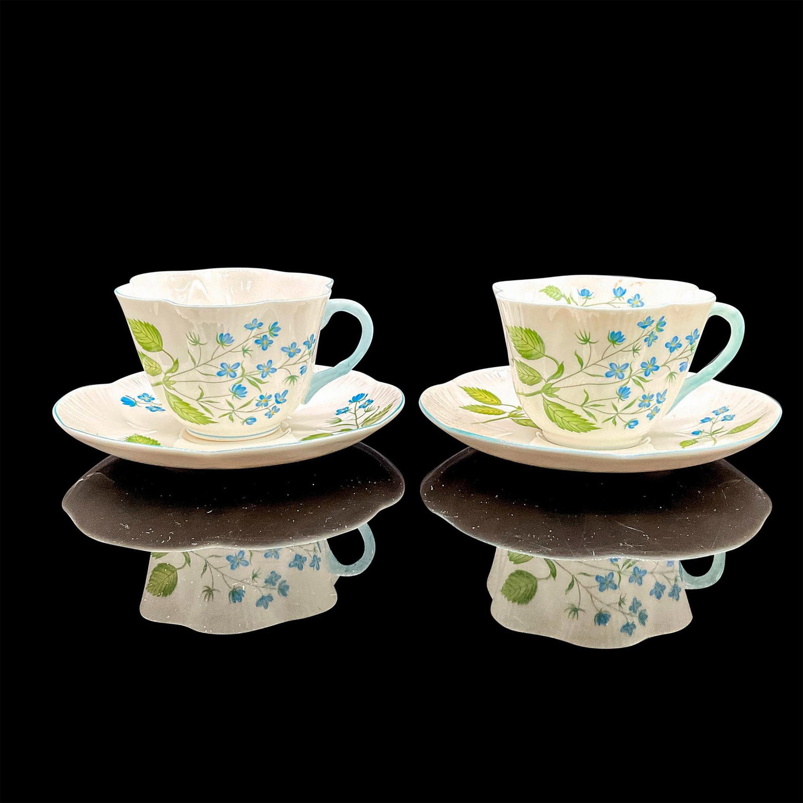 4pc Shelley England Cups and Saucers, American Brooklime: Vintage white bone china with delicate blue flowers, blue rims; two tea cups and two saucers. Dainty shape. Tea cups 4"L x 3.5"W x 2.5"H. Saucers 0.75"H x 5.5"dia. Shelley England backstamp. Manufactu