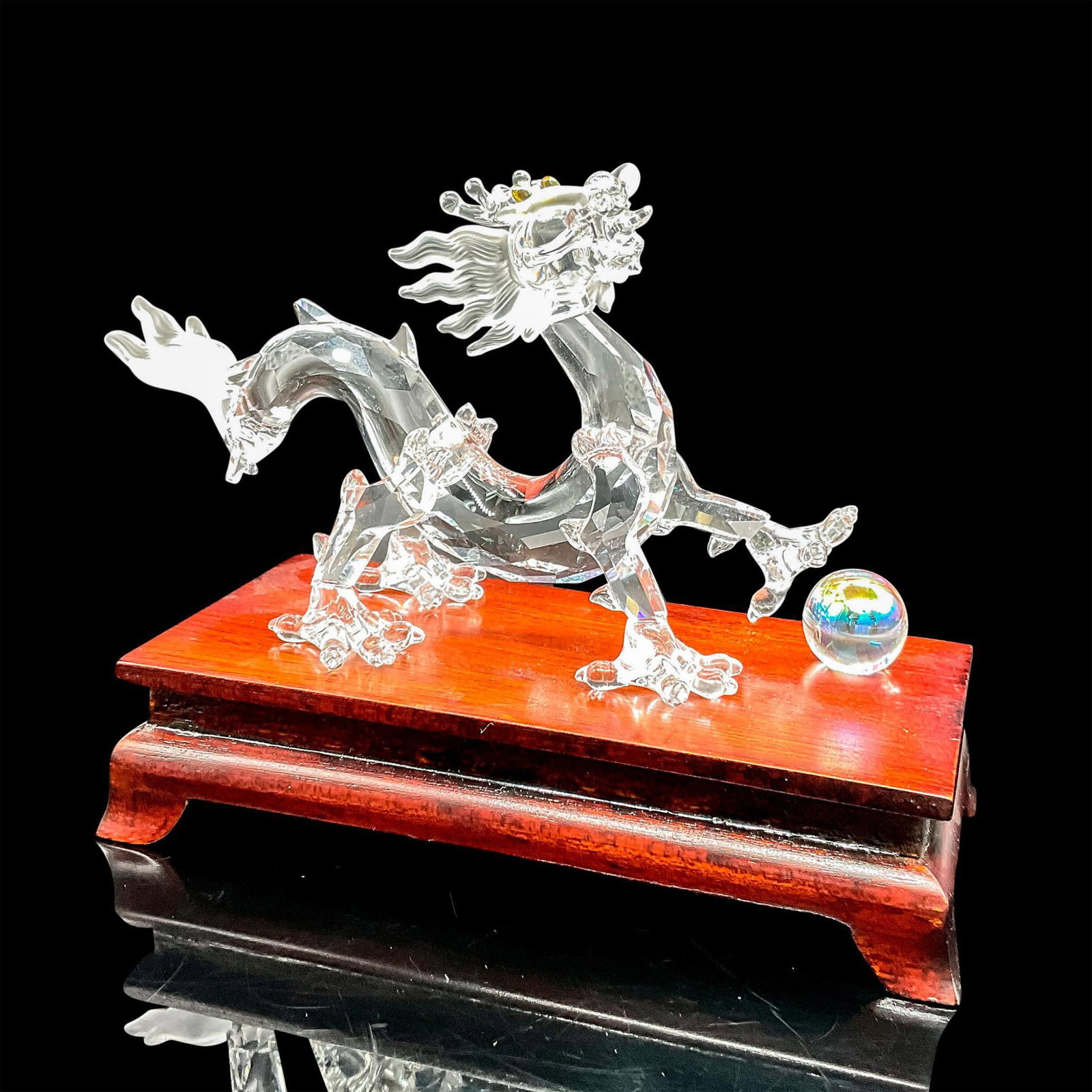 Swarovski Crystal Figurine + Wooden Base, Dragon 238202: Part of the Fables and Tales collection. Lead crystal dragon made from faceted clear crystal with frosted crystal details and crystal ball. Complemented with a beautifully polished teak wood stand. Sw