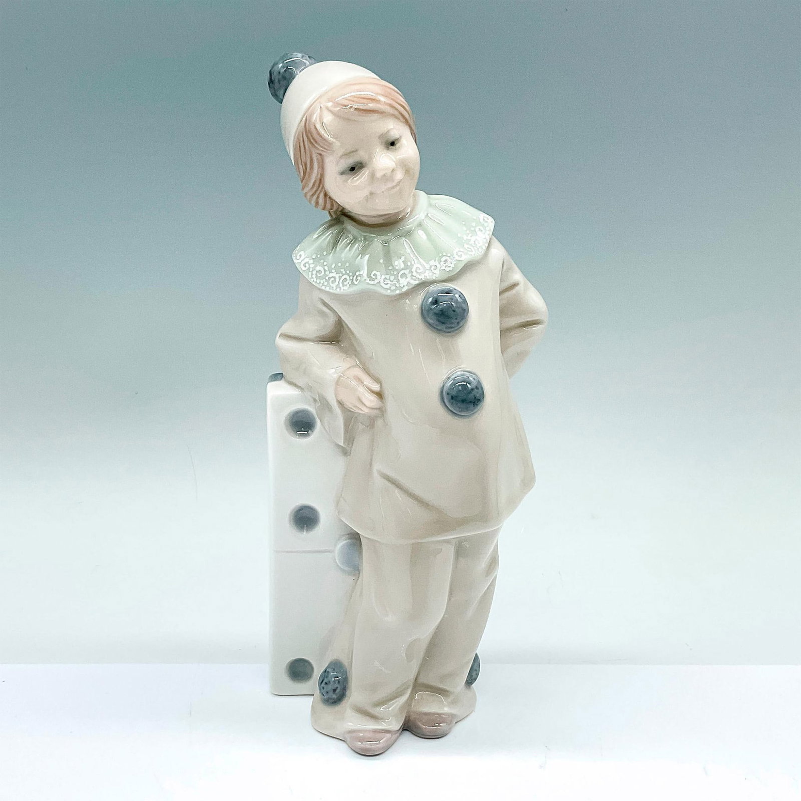 Girl with Domino 1001175 - Lladro Porcelain Figurine: Glazed figure of a young girl in a clown attire leaning against a large domino piece. Lladro backstamp. Artist: Juan Huerta Issued: c. 1971-1986 Dimensions: 3.5"L x 2.25"W x 7.75"H Manufacturer: Lladr