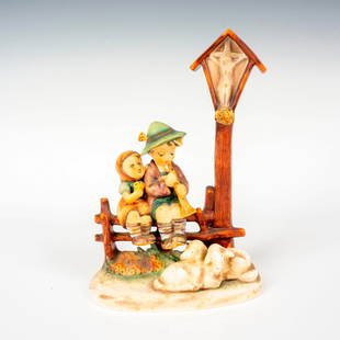 Goebel Hummel Figurine, Wayside Devotion: Hand painted in matte finish modeled as two kids sitting on a fence with two lambs resting by their feet. M. I. Hummel impressed backstamp. Goebel backstamp. Dimensions: 5"L x 2.5"W x 7.25"H Manufactu