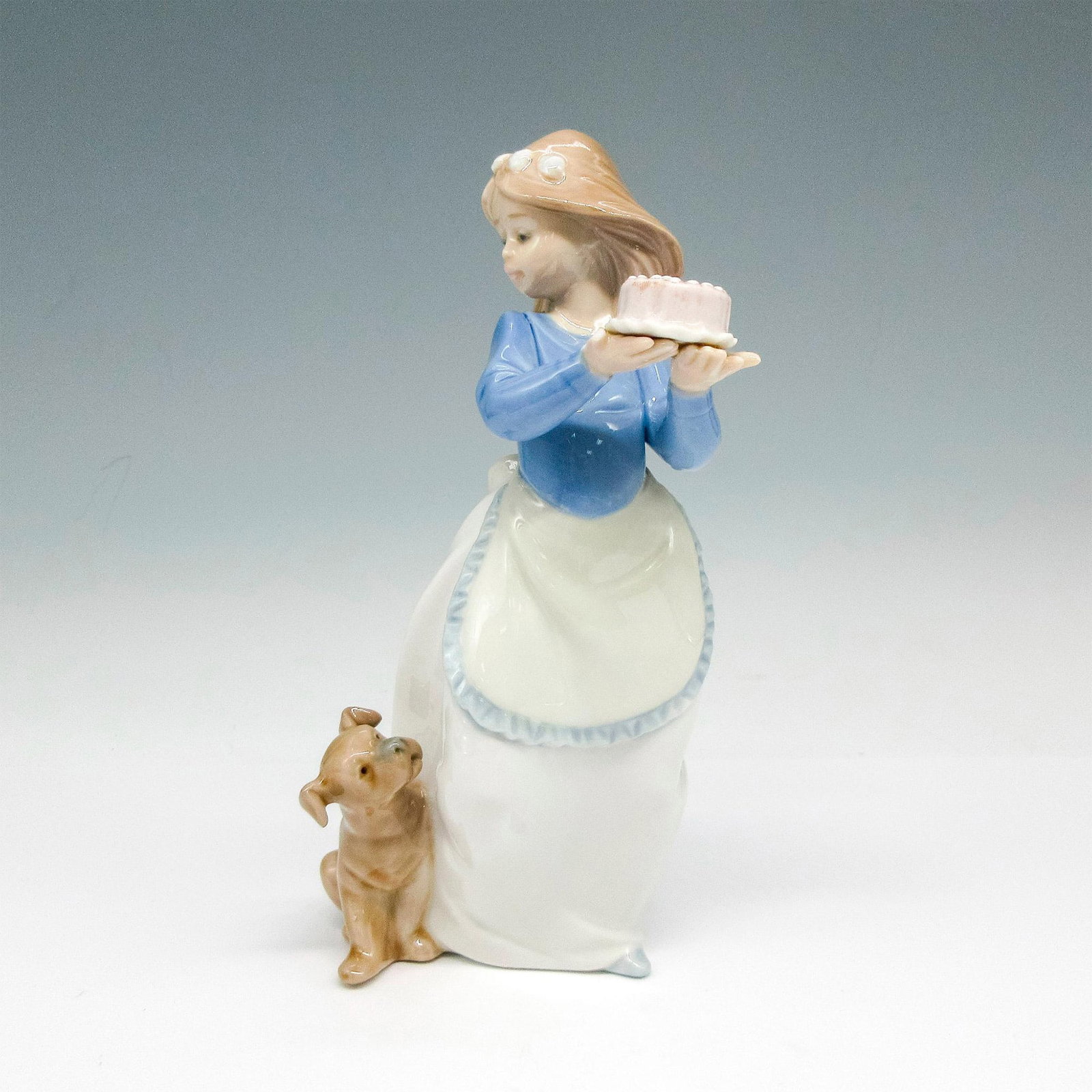 Puppy's Birthday - Nao by Lladro Porcelain Figurine: Glossy figural carrying a birthday cake with her dog looking up at her. Nao by Lladro backstamp. Artist: Regino Torrijos Issued: 2012 Dimensions: 3.25"L x 3.25"W x 7.5"H Manufacturer: Nao by Lladro C
