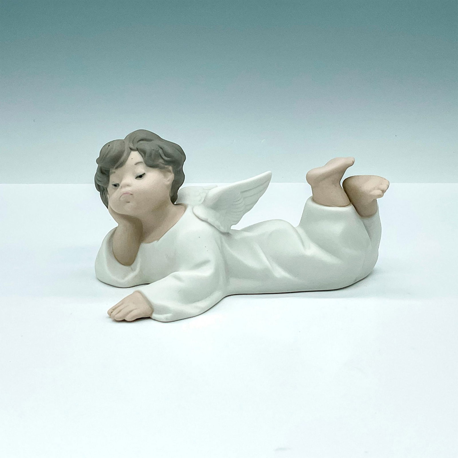 Lladro Porcelain Figurine, Angel Reclining 1014541: Captivating matte finished figurine of young angel laying on the floor with bored look on his cherub looking face. Crafted in subtle cream and brown hues. Lladro backstamp. Artist: Fulgencio Garcia Di