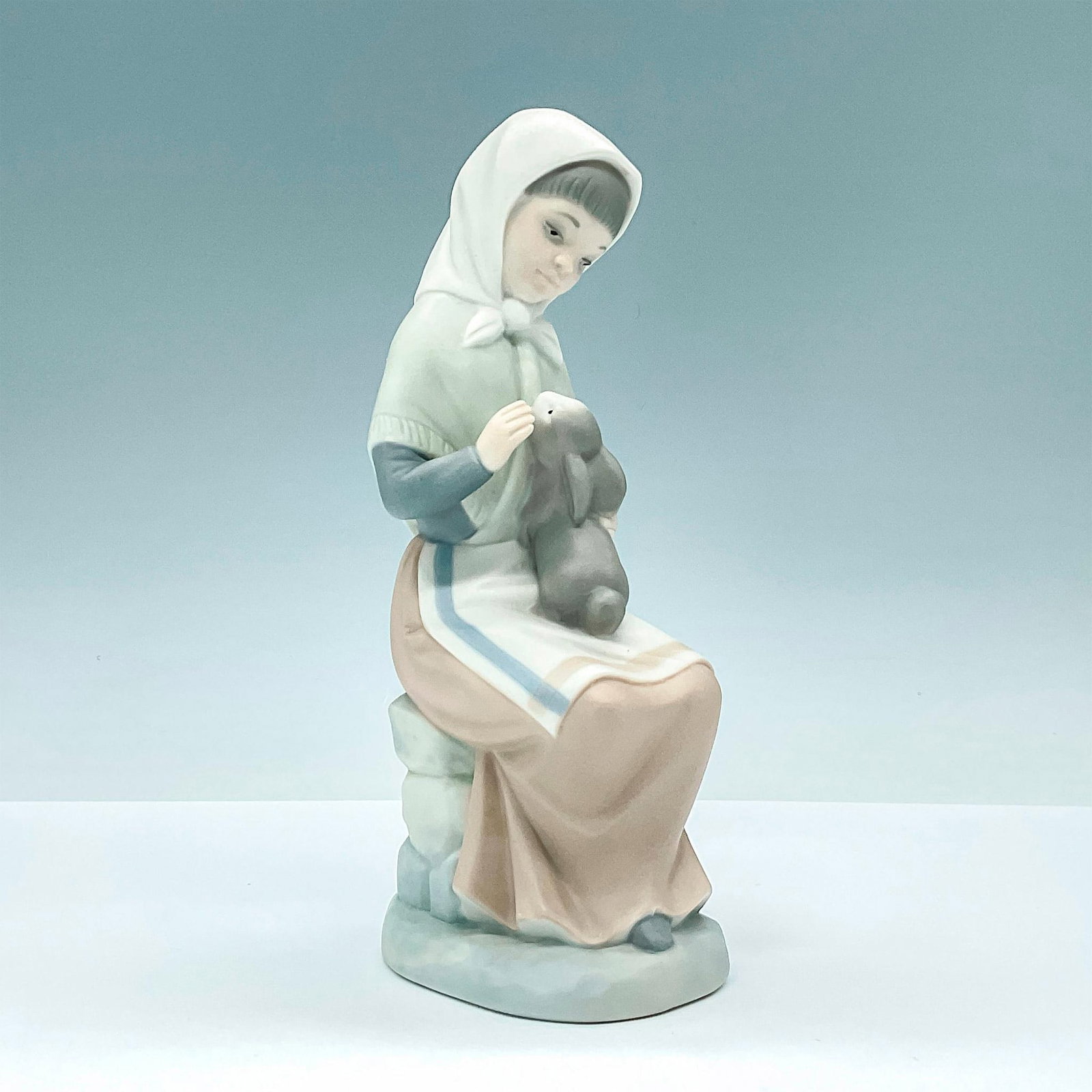Nao by Lladro Porcelain Figurine, Girl Holding Bunny: Lovely matte finished porcelain figurine of young woman holding a pet bunny on her lap. Crafted in subtle hues of blue and brown. Nao backstamp. Dimensions: 3.5"L x 3"W x 7.75"H Issued: 20th Century M