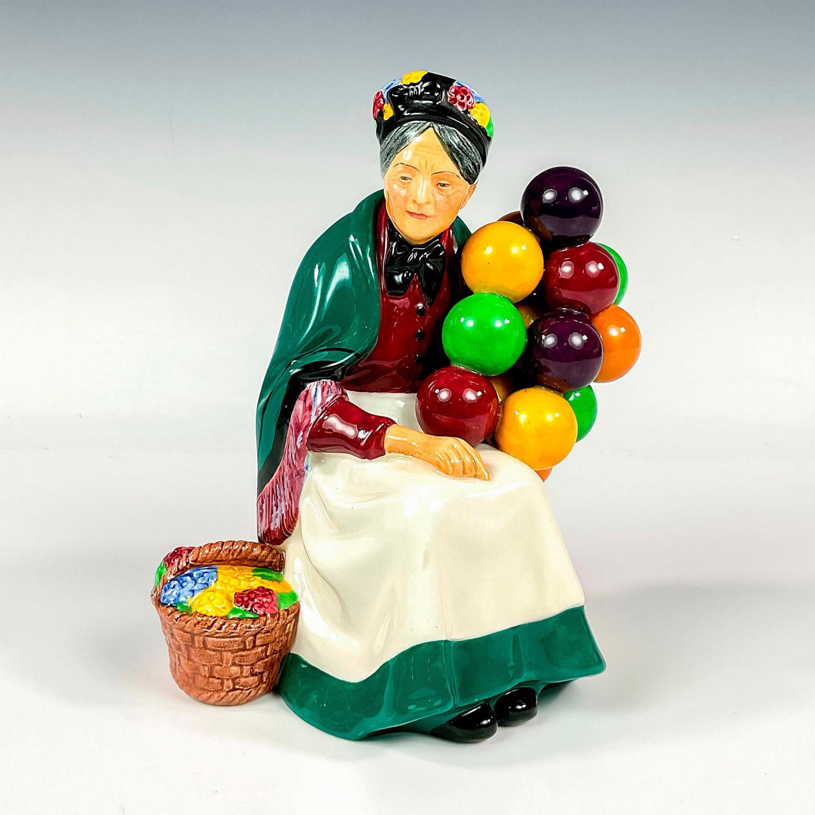Old Balloon Seller HN1315 - Royal Doulton Figurine: Glazed finish, modeled as an old lady selling balloons with a basket of flowers by her side. Royal Doulton backstamp. Artist: Leslie Harradine Dimensions: 5.75"L x 4"W x 7.5"H Issued: 1929-1998 Manufa