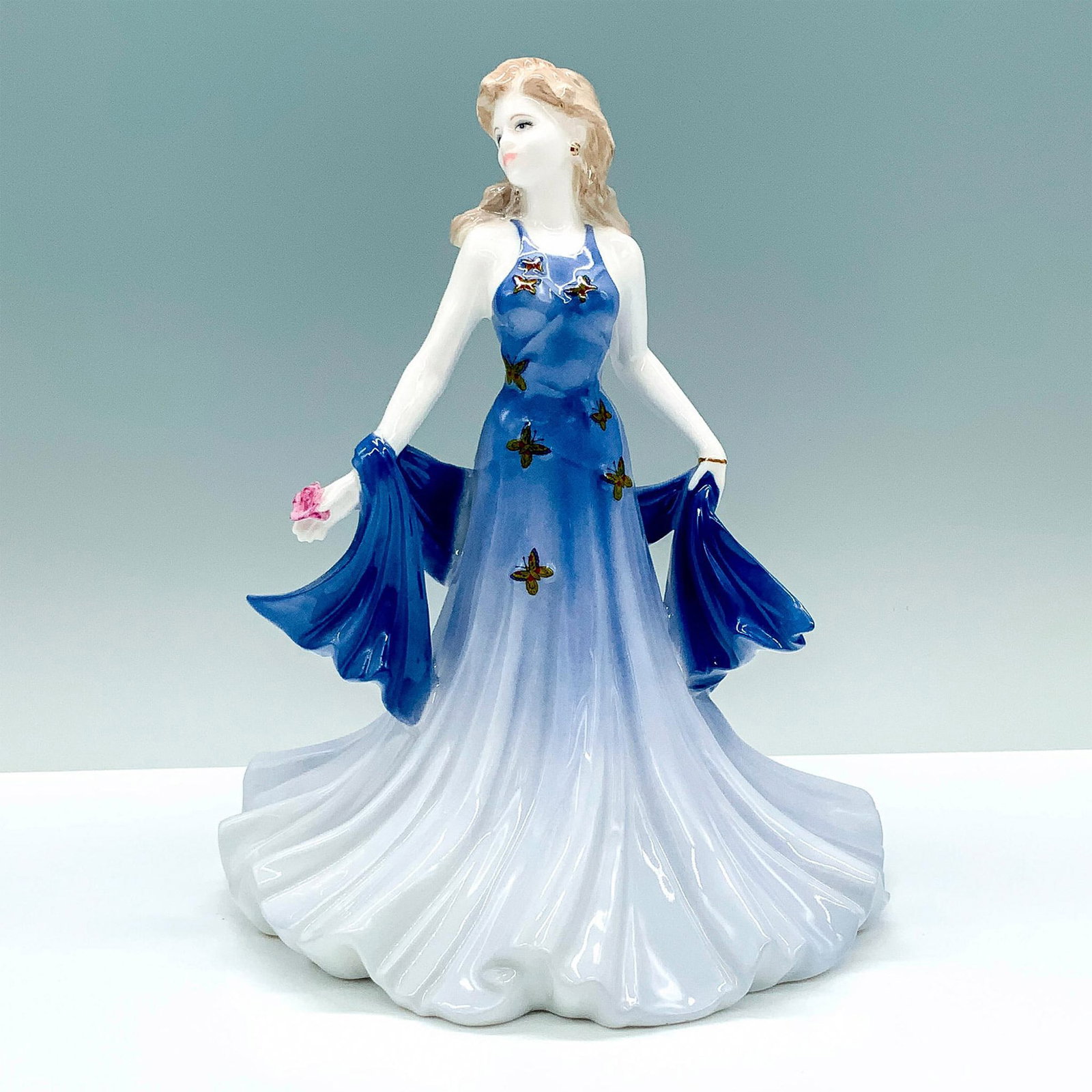 Coalport Ladies of Fashion Figurine, Shirley: A porcelain figure of a woman dressed in a dark blue dress that fades into lighter colors decorated with butterfly designs and holding a rose. Coalport backstamp. Artist: Jack Glynn Dimensions: 6.5"L 