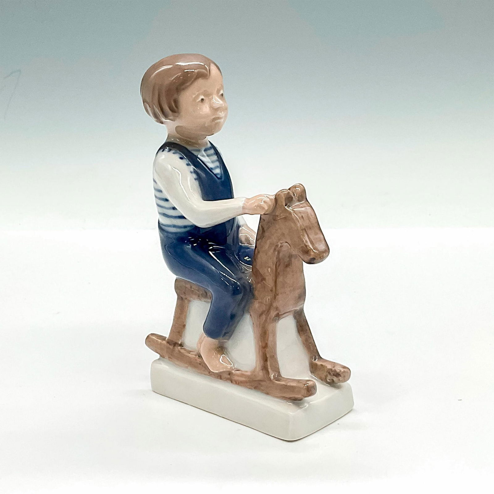 Royal Copenhagen Porcelain Figurine, Boy On Rocking Horse: Figure of a boy in overalls seated on a rocking horse. Over glazed finish. Royal Copenhagen backstamp. Issued: c. 1889 - 1922 Dimensions: 3.25"L x 1.5"W x 5.25"H Manufacturer: Royal Copenhagen Country