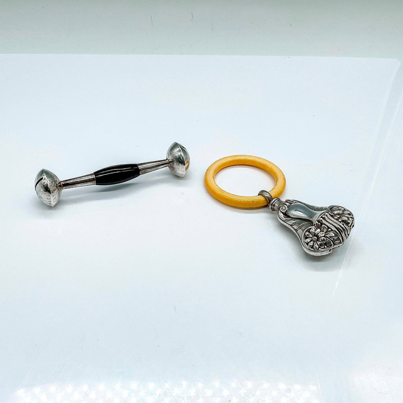 2pc Antique Silver Plated Baby Rattles: Two beautifully unique baby toys. One with a dark wooden handle. The second is a floral embossed rattle with a carved bone ring for teething. Rattle 1: 4"L x 1"W x 1"H. Rattle 2: 4.5"L x 2.25"W x 1"H.