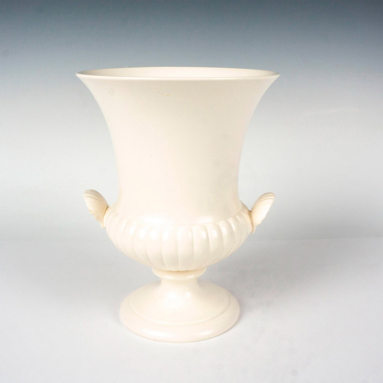 Wedgwood Medici Style Urn Vase With Shell Handles: This Wedgwood Moonstone Urn Vase is a beautifully crafted piece of Wedgwood Queen's Ware. It features a footed design with a ridged lower portion and twin shell handles that gracefully curve upwards. 