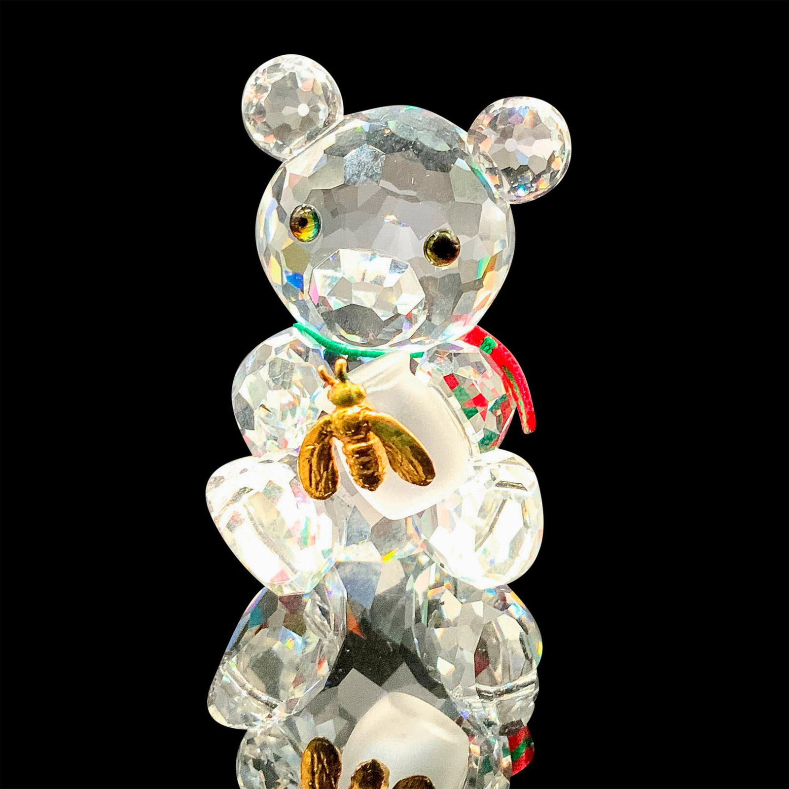 Swarovski Crystal Figurine, Kris Bear with Honey Pot: A clear crystal figure of a bear sitting down on their bottom while holding a honey pot that is decorated with a gold-colored bee. Melon colored gems are used for the eyes along with a green and red s